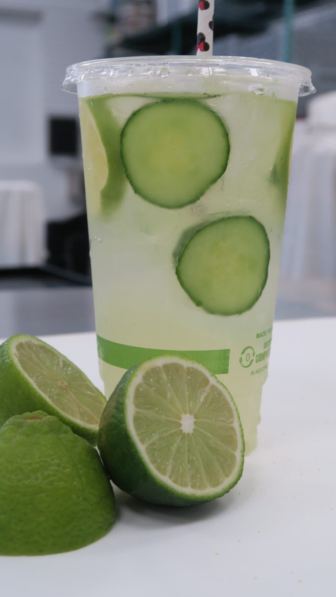 A clear cup of lime-infused drink with sliced cucumbers and lime halves nearby.